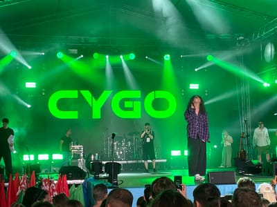 The image depicts a concert taking place outdoors, illuminated by numerous green stage lights that create beams and haze across the scene. In the background, a large, bright green LED screen displays the word "CYGO" in bold, capital letters. On stage, several individuals are visible. A lead singer, wearing a purple and black plaid shirt and black pants, stands center-stage, looking upwards with their mouth open. To the left of the singer, a man in a black t-shirt is walking across the stage, holding some papers. Behind him, a drum set and other musical equipment are set up. Another person, dressed in black, is standing near the drums, appearing to operate a camera. To the right of the lead singer, a woman in a white shirt and green pants is holding a microphone, and further to the right, a man in a white shirt and shorts is standing. In the foreground, a crowd of people is visible, some holding up cell phones to take pictures or record. Red flags and green fabric are arranged along the front of the stage, possibly as decoration or barriers. The concert appears to be part of an event at an outdoor venue. The presence of the "CYGO" logo strongly suggests this is a performance by the artist of that name. Given the context of concerts and public performances, this scene is likely located in a city park, festival grounds, or stadium within the city of Brest, Belarus.
