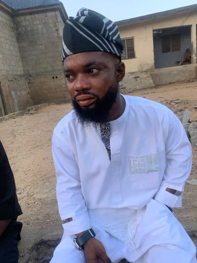 The image portrays a male individual in Okene, Nigeria, positioned outdoors under daylight conditions. The subject is a young adult male with dark skin, a short dark beard, and a trimmed haircut. He wears a black and white striped traditional cap and a long-sleeved white tunic, which features a black and white patterned fabric at the neck opening and light-colored, possibly beaded, embellishments forming a design on the left chest pocket and along the lower portion of his trousers. A dark-strapped smartwatch is present on his left wrist. He is looking towards the left side of the frame with a neutral expression.
To the immediate left, a partial view of another person, clad in a black garment, is visible. The immediate ground consists of dry, light brown soil with scattered small rocks and debris. In the background, two structures are visible: on the left, an unpainted concrete block building with a dark corrugated metal roof; on the right, a single-story building with a faded light-colored exterior wall, a window, and a door. A distant, indistinct individual is seated on a bench outside the right-side building.