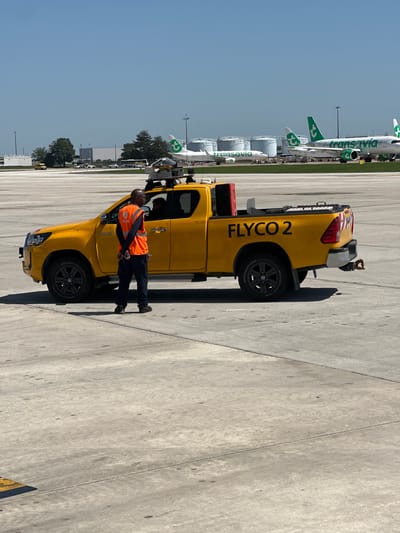The image shows an outdoor scene at the airport in Paray-Vieille-Poste, France. A yellow pickup truck labeled "FLYCO 2" is parked on a concrete apron. A man wearing an orange reflective vest is standing near the truck. In the background, several airplanes with green and white livery, one labeled "Transavia," are visible along with other airport infrastructure. The sky is clear and blue.