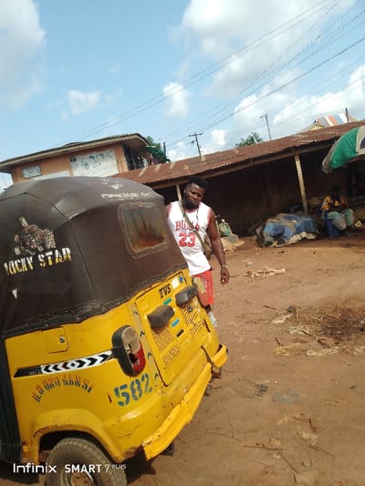 A yellow "Lucky Star" branded auto rickshaw or "keke" sits on a dirt road. The vehicle has blue lettering and the number 582 on its side. Next to it, a man wearing a Bulls jersey and red shorts is walking. In the background, a rusty-roofed building houses some people and goods. Utility poles and wires stretch overhead against a partly cloudy sky. The image appears to have been taken with a "Infinix SMART 7 PLUS" phone, as printed in the lower left corner.