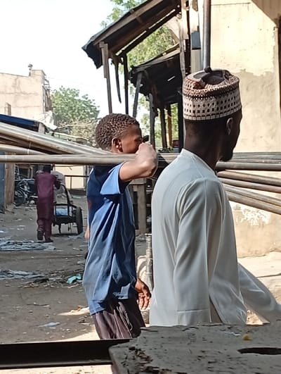 The image depicts an outdoor scene in Kofar Ruwa, Nigeria, under clear daylight conditions. In the immediate foreground, two dark-skinned males are engaged in transporting long, silver-colored metal rods. The individual on the left, wearing a blue t-shirt and dark trousers, carries multiple rods across his right shoulder, using his raised left hand to steady them. To his right, a second male, partially visible, wears a white long-sleeved garment and a patterned brown and white kufi cap on his head, walking parallel to the rods. The ground beneath them is an uneven dirt path littered with miscellaneous debris. In the mid-ground, a third dark-skinned male, dressed in a maroon long-sleeved shirt and trousers, pushes a two-wheeled metal handcart away from the camera. Behind these individuals, dilapidated wooden structures with corrugated metal roofs are visible. The background on the left features a multi-story, light-colored building with a flat roof, while sparse green foliage is present towards the upper right.
