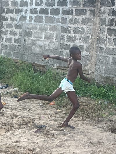 A young boy is performing a handstand on a sandy area next to a grassy patch and a concrete wall. He is wearing white shorts. The sandy area appears to have some scattered debris. The concrete wall is made of rough-hewn blocks. The scene is outdoors, and based on the lighting, it appears to be daytime. The boy's body is angled upwards, with his legs extended in the air. His face is turned downwards, and he seems focused on his balance. The overall impression is of a playful or athletic activity. The text "GRANGE" is visible on the waistband of the boy's shorts. The location is Badagry, Nigeria.