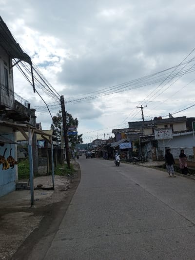 The image captures a street scene, identified as being in Ariyalai, Sri Lanka, although prominent signs, such as one reading "PRAKTEK DOKTER ASYFA", display Indonesian language text. The scene is set under an overcast sky with dense grey clouds. An asphalt road, appearing dry, leads into the distance with a slight curve to the left. Numerous overhead utility lines crisscross the frame, supported by dark utility poles positioned along both sides of the street.
On the far left foreground, an older, light-colored multi-story building with a balcony is partially visible, from which multiple black cables descend. Adjacent to it, a light blue stall with a corrugated roof features several bunches of bananas hanging from a crossbeam. Behind this, a prominent dark utility pole stands with attached signage. Further down the left side, various buildings and some vegetation are observed.
On the right side of the road, two women are standing on a paved shoulder or sidewalk next to a drainage ditch. The woman closer to the foreground wears a dark top and a light-colored patterned skirt. The woman behind her wears a head covering and a patterned dress or tunic. Behind them, a series of single and multi-story buildings line the street. A significant white rectangular sign with red text, "PRAKTEK DOKTER ASYFA", is mounted on a building. A white scooter, carrying a single rider, is in motion, traveling away from the viewer on the right side of the road. In the mid-ground and background, the street continues with more buildings, utility poles, and a dark vehicle visible further in the distance.