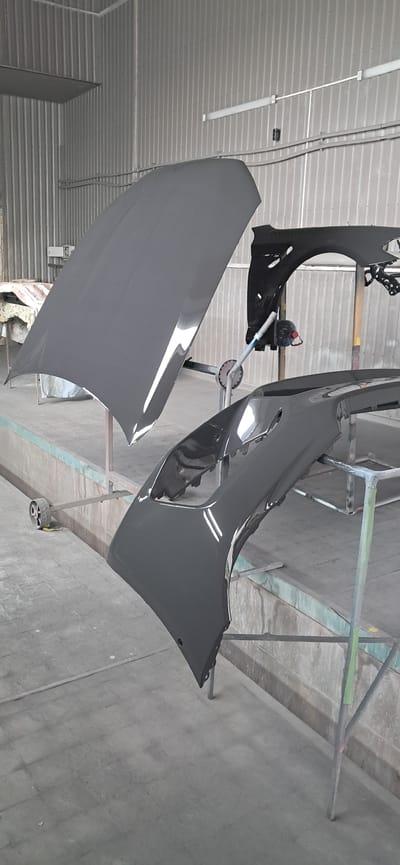 In a workshop, likely a car repair shop, located in Horishni Plavni, Ukraine, a car's hood and bumper are displayed on stands. The hood is open and angled upwards, while the bumper is positioned lower down. Both car parts are painted a dark grey color and appear to be freshly finished or in the process of being painted. The workshop environment features a grey corrugated metal wall in the background and a concrete floor. The lighting suggests an indoor setting, possibly with artificial lights present, as indicated by reflections on the car parts. There are no people visible in the image, and no specific actions or interactions are taking place. The scene is static, focusing solely on the automotive components.