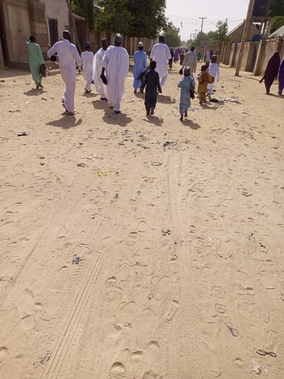 The image shows a group of people walking on a dirt road in Maiduguri, Nigeria. The road is covered in sand and has tire tracks. The people are all wearing traditional clothing and walking in the same direction. The environment is dry and dusty. The scene is captured from a high angle, looking down on the group as they walk away from the viewer. There is a building in the background to the left of the image. There are no trees to the right of the image, only a wall. The road is empty except for the people in the image, with some debris scattered on the ground.