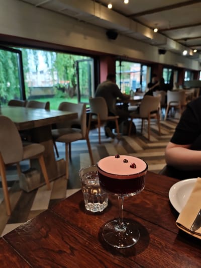 A photograph taken indoors depicts a restaurant scene. A stemmed cocktail glass filled with a dark liquid and topped with a light pink foam and a few small dark garnishes is on a dark wooden table, next to a glass of water and a plate with cutlery. In the blurred background, several people are seated at tables, and there is a view through large windows of outdoor greenery. The location appears to be a restaurant in Vinnytsia, Ukraine.