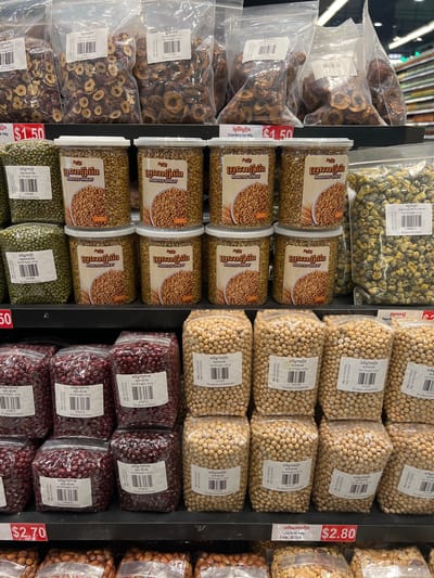 The image shows a shelf display of dried food products in what appears to be a market or grocery store in Siem Reap, Cambodia. The products are packaged in plastic bags and containers and include dried red berries, green beans, roasted wheat, dried flowers, red beans, and soybeans. Some of the packaging features labels with text in Khmer script and English. Price tags are visible, with one indicating "$2.70" and another "$2.80". The overall environment suggests a commercial setting with multiple shelves stocked with goods. There are no people visible in the image, and the focus is solely on the product display.