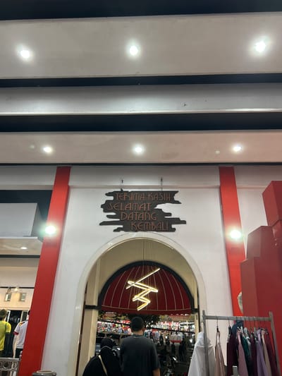 The image shows the entrance to a marketplace or shopping area in Depok, Indonesia. Above the arched entrance, a dark wooden sign with carved text reads "TERIMA KASIH SELAMAT DATANG KEMBALI," which translates to "Thank you, welcome back." The entrance is flanked by red pillars and white walls, illuminated by ceiling lights. Inside the arch, the marketplace is visible, with rows of stalls displaying clothing and other goods, and people walking around. A modern, geometric light fixture hangs from the ceiling within the archway. To the right of the entrance, a rack of colorful clothing is displayed for sale. The overall scene suggests a lively commercial environment.