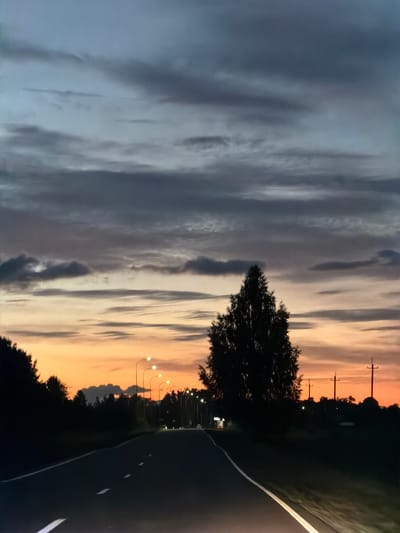 The image depicts a desolate, paved road stretching into the distance, flanked by dark silhouettes of trees and vegetation. The sky overhead is a dramatic canvas of twilight hues, with streaks of orange and yellow from the setting sun blending into a gradient of soft blues and grays, overlaid with textured clouds. Streetlights dot the roadside, their illumination casting a faint glow. A lone, large tree stands prominently on the right side of the road, its dark form silhouetted against the vibrant sky. In the far distance, there are indications of more lights, suggesting a settlement or infrastructure, possibly including utility poles. The overall atmosphere is one of quiet transition from day to night in a rural or semi-rural setting, with no people or animals visible. The scene does not contain any elements that would allow for a specific location within the city of Крынды, Russia to be identified.