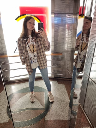 A young woman captures a selfie in what appears to be an elevator or a modern interior space in Al Jizah, Jordan, characterized by numerous reflective surfaces and intricate architectural details. She stands upright, holding a dark-colored smartphone to take the picture. She has long, dark brown hair and a calm, pleasant expression. She is dressed in a brown and beige plaid shirt worn over a light t-shirt featuring a stylized sun graphic. Around her neck is a lanyard with multiple "Deloitte" logos printed on it, to which an ID card is attached. She wears blue jeans and light-colored sneakers with thick laces.
The setting is highly reflective, with mirrored panels on the walls and ceiling that create multiple distorted reflections of the woman and the interior. The walls are composed of textured, metallic panels, while the floor features an elaborate geometric pattern of various speckled stone tiles—including light grey, reddish-brown, and dark greenish-grey—outlined by brass or gold-colored inlays. Bright light emanates from the left, casting reflections and highlights across the polished surfaces. The overall impression is one of a sleek, contemporary, and well-lit indoor environment, likely during daytime.
