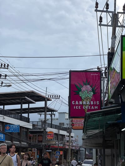 The image shows a street scene in Karon, Thailand, with a focus on the signage and buildings. The orientation of the image is rotated 90 degrees counterclockwise, making it appear vertical. Overhead, a complex network of electrical wires and poles crisscrosses the sky, which is overcast and suggests a cloudy or dull day.

Prominently featured is a bright pink sign advertising "CANNABIS ICE CREAM" with an illustration of an ice cream cone adorned with cannabis leaves. Below this, several Burger King signs are visible, one with the slogan "FLAME GRILLING SINCE 1954." A pharmacy sign is also present, written in Thai and English, with the word "PHARMACY" clearly legible.

The street level shows glimpses of shops and businesses, along with several people. Some individuals are walking or standing, while others appear to be sitting or riding a vehicle. The expressions on the faces of those clearly visible are neutral. The overall impression is of a busy commercial street, characteristic of a tourist area. The time of day is not precisely discernible due to the overcast sky, but it appears to be daytime.