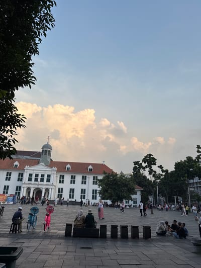 The image depicts the Fatahillah Square in the Old Town of Jakarta, West Jakarta, Indonesia. In the foreground, several people are gathered. To the left, a man wearing a white cap and blue shirt sits on a stool beside a blue bicycle. Next to him is a pink bicycle. Further to the right, two individuals, one in a light brown hijab and the other in a dark green hijab, are seated on a dark bench. A group of people are also seen sitting on the ground in the middle right of the frame, engaged in conversation. In the background, the historic white building, the Jakarta History Museum (formerly the City Hall), stands prominently with its red tiled roof. People are seen walking and congregating in front of the museum. Trees are interspersed throughout the scene, with a large tree branch in the upper left corner partially obscuring the sky. The sky is partly cloudy, with large, soft clouds illuminated by sunlight.