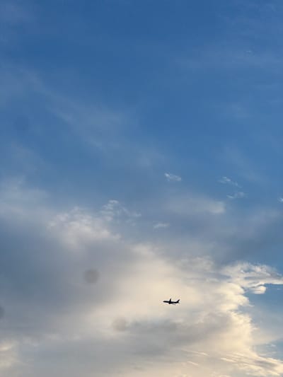 An airplane is flying in the sky over Antalya, Turkey. The sky is blue with scattered clouds. The airplane is a silhouette against the clouds. The image is taken from a low angle, looking up at the airplane.