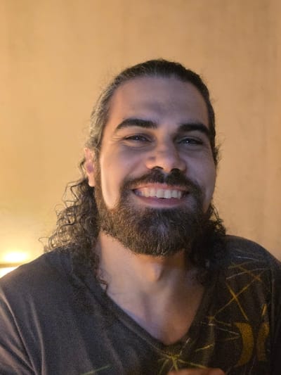 A close-up portrait photograph depicts an adult male situated indoors in Villa San Andrés, Argentina. The subject features dark, curly hair pulled back, interspersed with grey strands, and a full, dark beard and mustache. He is smiling, exposing his upper teeth, and directs his gaze towards the camera. He wears a dark-colored V-neck t-shirt, which displays a yellow-gold geometric pattern and a partial 'Hurley' logo on the left chest area from the viewer's perspective. The background is an out-of-focus, plain, light orange wall, with a warm light source visible in the lower-left portion of the frame.