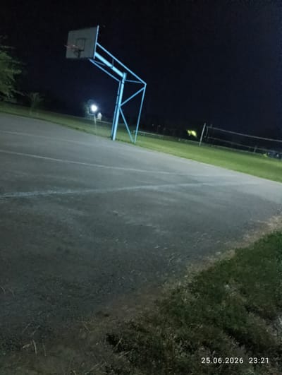 The image depicts an outdoor basketball court at night, illuminated by artificial lights. A blue basketball hoop with a backboard and rim stands prominently on the left side of the court. The court itself is paved and has white lines marking its boundaries. Adjacent to the court, to the right, is a grassy area with a volleyball net visible in the background, suggesting a multi-purpose recreational space.

The lighting creates a dark ambiance, with the basketball hoop and court being the most illuminated elements. A few distant lights are also visible, possibly streetlights or lights from nearby buildings. The time stamp "25.06.2026 23:21" indicates the photo was taken late in the evening.

There are no people visible in the image, and the scene appears to be deserted. The weather is not discernible from the image. The presence of both a basketball hoop and a volleyball net hints at a public park or sports facility in a community setting. The overall mood is quiet and still, characteristic of a nighttime scene with no activity.