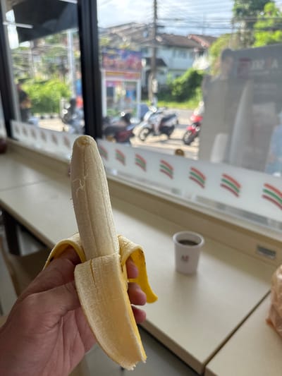 A person's hand holds a partially peeled banana in the foreground. Behind the banana, through a window, a street scene in Rawai, Thailand, is visible. Several motorcycles are parked on the street, and buildings with signs are visible in the background. A small white cup containing a dark liquid is on a counter to the right of the banana. The scene appears to be inside a convenience store.