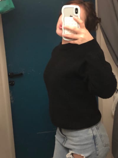 A person holds an iPhone in a mirror selfie. They're wearing a black sweater and distressed light-wash jeans. The background features a blue wall, a white baseboard, and a coat hook. The lighting is somewhat dim. The image is rotated counterclockwise.