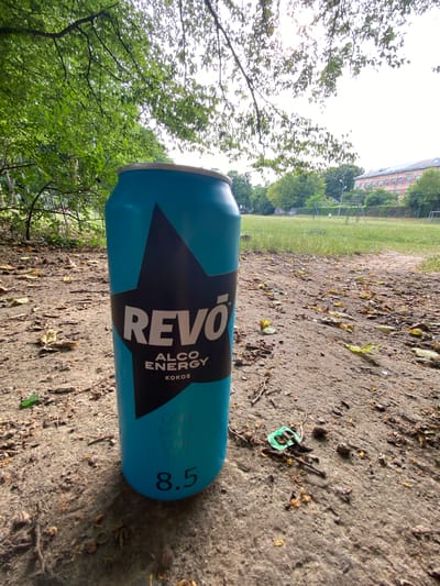 An upright, blue aluminum can of "REVO ENERGY" lies on its side on a dirt path with scattered leaves and debris. The can features a large black star with the word "REVO" in bold white letters across it. Below "REVO," smaller white text reads "ALCO ENERGY KOKOS." To the right of the star, there are vertical lines of text that appear to read "ONE SHOT ONE HIT" and a large "8.5" is printed on the right side of the can.
In the background, a grassy field borders a tree line. Further back, a building with multiple windows is visible. The scene appears to be outdoors, possibly in a park or a recreational area. A small, green plastic object, possibly a bottle cap or tab, is visible on the dirt path near the can. The lighting suggests it is daytime, with the sky appearing bright and possibly overcast. The image's orientation is rotated, with the can lying horizontally and the background elements tilted. There are no people visible in the image.