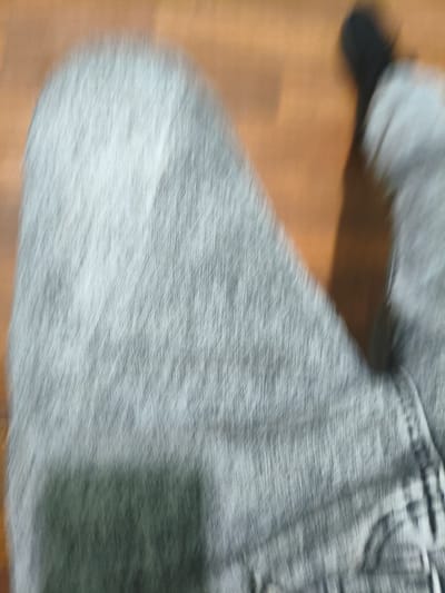 This is a very blurry, abstract, close-up shot of someone's legs and feet, likely taken from a low angle. The main subject is a person wearing light-colored, textured pants, possibly denim, which are the dominant feature of the image. The texture of the fabric is emphasized by the motion blur, creating streaks of light and shadow that give the pants a dynamic appearance. One foot is visible at the top right of the frame, wearing a dark shoe or sock. The background is a blurry expanse of warm, orange-brown tones, suggesting a wooden floor or a similar surface. The overall impression is one of movement, as indicated by the strong motion blur throughout the image. There are no specific details about the time of day, weather, or emotions, but the blur suggests an candid or accidental capture during movement. No text is visible in the image.