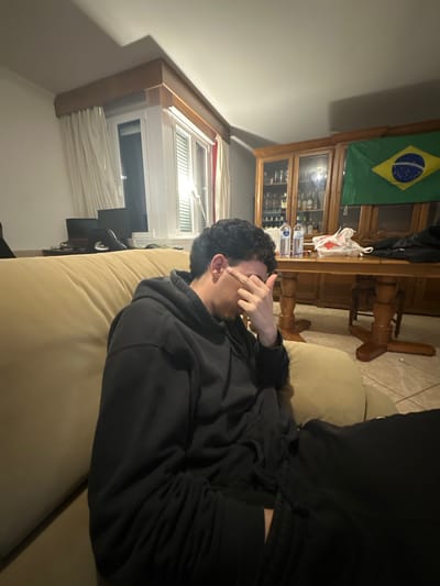 A person with dark, curly hair lies on a light-colored sofa, covering their eyes with a hand. They are wearing a dark hooded sweatshirt and dark pants. The room contains a wooden cabinet displaying bottles and glassware, topped with a Brazilian flag. A wooden table with decorative legs stands near the cabinet. A window with white blinds is visible in the background, adjacent to a dark-colored object, possibly a television or monitor. The location is likely indoors, potentially in a home. I cannot confirm this is Bragança, Portugal.
