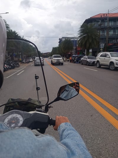 Captured from the perspective of a person riding a scooter, the image shows a street in Ban Ao Nang, Thailand. The rider is wearing a light blue denim jacket and a helmet, visible on the left. The scooter features a speedometer with a Honda logo, reading 61615 km. A camouflage bag is mounted near the front of the scooter.

The road has two yellow lines indicating traffic lanes. Traffic includes cars (a white pickup truck, a grey sedan, and a white SUV), parked motorcycles, and pedestrians on the side of the road. Buildings, including a multi-story structure with a red roof and palm trees, line the street. The sky is overcast. The scooter's side mirror reflects the rider taking the picture with their phone.
