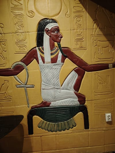 The image displays a detailed bas-relief carving of an ancient Egyptian figure, likely a pharaoh or deity, seated on a throne. The figure has dark skin, black hair with white streaks, and is adorned with a white kilt-like garment and a white headdress. They are holding an ankh, a symbol of life, in their left hand, and their right arm is extended. Hieroglyphics and other Egyptian motifs are carved into the surrounding wall. The carving is rendered in a warm, yellowish-brown tone, with accents of white, black, and dark green. In the lower right corner of the image, a modern electrical outlet is visible, incongruously integrated into the ancient-themed wall.

This bas-relief is located in the Museum of Aviation in Smolyan, Bulgaria.