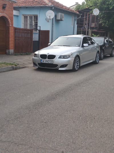 This image depicts a residential street scene in the city of Lom, Bulgaria. The foreground features an asphalt road with two parked vehicles on the right side.
The primary subject is a silver BMW 5 Series sedan (E60 generation) parked parallel to the curb, facing towards the left. It has a sunroof, multi-spoke alloy wheels, and a black kidney grille. The front license plate displays "BG M 6942 CE". Behind the BMW, partially obscured, is a black Volkswagen Golf Mk4 hatchback, also parked.
To the left of the vehicles, a concrete sidewalk or paved area borders a light blue single-story house with a red tiled roof. A window with white frames and metal security bars is visible on the house's facade. Mounted on the house's exterior are a white satellite dish with "Булсатком" (Bulsatcom) branding and an outdoor air conditioning unit. A brown wooden fence with a small black metal gate, marked with the number "15", encloses the property. To the far left, a brick archway is partially visible.
The ground consists of asphalt pavement for the road and concrete for the sidewalk, with sparse vegetation and weeds growing in cracks. Green foliage from trees is visible in the background above the buildings. No people are present in the image. The scene is illuminated by diffused daylight, suggesting an overcast sky or soft lighting conditions.