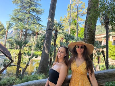 The image captures two women in a natural, outdoor setting with tropical vegetation. The woman on the left is wearing a black strapless top, while the woman on the right is wearing a yellow floral dress and a wide-brimmed straw hat with sunglasses. They are leaning against a low stone wall or barrier. In the background, there is a small lake or pond, with various palm trees and other greenery surrounding the water. The trees include palm trees and taller, broad-leafed trees. Behind them, there are structures covered with green vines, suggesting the presence of buildings. The image was taken in Bunyola, Spain.