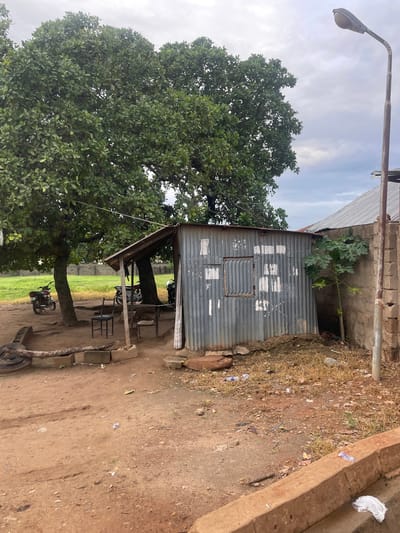 The image depicts an outdoor scene in Keffi, Nigeria, featuring a small, weathered corrugated metal structure with a wooden roof. The structure appears to be a makeshift shop or shelter, with a window covered by a metal grate. In front of it, two chairs and a table are set up on a dirt ground. A motorbike is parked to the left of the structure, near a large tree. Another motorbike is visible further back, partly obscured by the trees. A tall streetlight stands to the right of the metal building, next to a concrete wall. The ground is covered in dirt, dry grass, and scattered debris, including plastic bottles and bags. In the background, a grassy field is bordered by a low brick wall, and additional structures with corrugated metal roofs are partially visible. The sky is overcast.