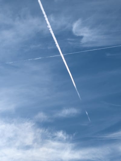 A bright blue sky with wispy cirrus clouds is bisected by two contrails. One contrail is thick and spans diagonally across the image. The other contrail is thin and vertical, intersecting the thicker one. The clouds vary in density, with some areas appearing almost clear. The contrails are crisp and white, contrasting with the blue sky.
