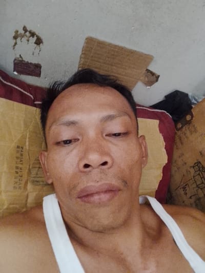 The image is a selfie taken by a man lying down, appearing to be indoors. The man has short dark hair and is wearing a white tank top. He has an expression of mild disinterest. The background is cluttered with various objects, including cardboard boxes and what appears to be a mattress or cushions. There is also some damage or debris on the ceiling in the upper left corner of the image. The overall impression is that the person is in a modest or possibly disheveled living space. There is no visual information in the image that allows for the specific identification of the location as Bukit Ketok, Indonesia.