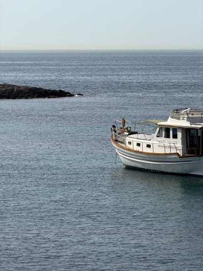 A white motor yacht named "MARLIN" is anchored in the calm, blue waters off the coast of Calvià, Spain. The yacht has a wooden trim and several windows. In the background, a rocky outcrop emerges from the sea. The sky is a pale, hazy blue. The overall scene depicts a serene coastal landscape.