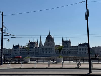 This media file captures a daytime scene in Budapest, Hungary, featuring the iconic Hungarian Parliament Building across the Danube River. The Parliament Building, a grand Gothic Revival structure with multiple spires and a prominent dome, dominates the background. In the foreground, a street or promenade runs along the riverbank.
Visible elements in the foreground include a street with a road and a pedestrian/bike path marked with red and pink lines. Several metal bike racks are present, with a bicycle parked in one of them on the right side of the image. A bench is also visible, with two people standing near it on the far right, seemingly taking in the view. Electrical poles with wires for overhead transportation systems, likely trams, are also a significant feature of the foreground and midground, stretching across the frame. Planters with small trees are positioned along the barrier separating the promenade from the river.
The weather appears to be clear and sunny, with a bright blue sky. The time of day is likely mid-morning or early afternoon, indicated by the strong sunlight and the shadows cast. The overall mood is one of urban tranquility and civic pride, with the magnificent Parliament Building serving as a majestic backdrop. No visible text is present in the image.