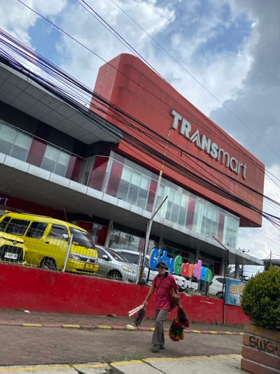 The image depicts a storefront of a large department store in Cimahi, Indonesia, located at coordinates -6.869, 107.534. The store is marked with a large sign reading "Transmart" in white lettering against a red background. The building has large windows, and a red awning above. To the left of the building, a yellow minivan is parked near a red brick wall. There is a man walking on the sidewalk in front of the store, carrying a brown satchel and some colorful objects. The man is wearing a red shirt. The sidewalk has yellow stripes painted on it. The sky is blue with white clouds. The ground is paved with concrete and cobblestones. There are many power lines crisscrossing the sky. There is a sign on the side of the building that says "City Garden". There is a green bush in the foreground.