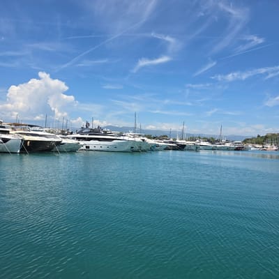 The image depicts a serene view of a marina in Antibes, France. In the foreground, the calm, turquoise waters of the Mediterranean Sea gently lap against the shore, reflecting the bright blue sky and scattered white clouds. A line of large, modern yachts are moored in the middle ground, their sleek white and dark hulls gleaming in the sunlight. The yachts are docked side-by-side, creating a striking display of maritime luxury. In the background, a lush green coastline rises, dotted with trees and distant hills, suggesting the picturesque natural beauty of the French Riviera. The sky is a vibrant blue, with wispy white clouds and faint contrails from airplanes stretching across it, indicating a clear and pleasant day. No people are visible in the image.