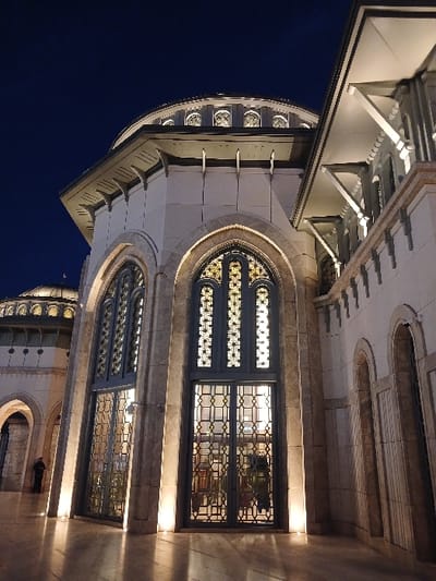 The image shows a portion of the exterior of the Camlica Mosque in Istanbul, Turkey. The mosque is a large, modern structure with a white facade and ornate detailing. The scene is captured at night and the mosque is illuminated, showcasing its intricate architectural details, particularly the large arched doorway with intricately patterned iron gates. The doors are closed and no people are visible. The image focuses on the mosque's facade, emphasizing its grandeur and architectural beauty.