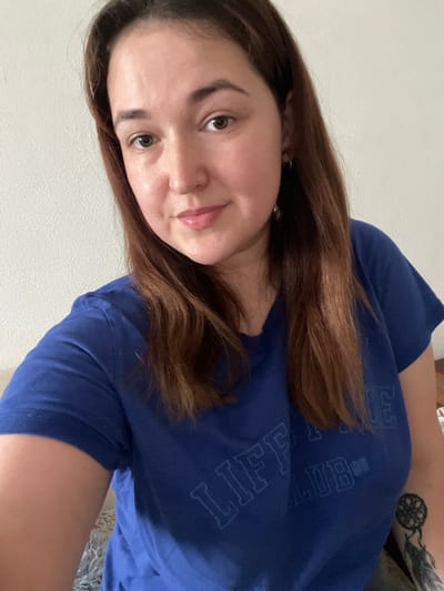 A woman is taking a selfie in front of a white textured wall. She has medium-length, reddish-brown hair and is wearing a blue t-shirt with the words "LIFE CLUB '96" printed on it. She has a dreamcatcher tattoo on her right arm. The image appears to be taken indoors, likely in a residential setting. There is no information available to indicate the location in Chelyabinsk, Russia.