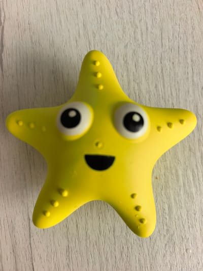A yellow rubber starfish toy with a happy face is positioned on a light-colored, textured wooden surface. The starfish has large, circular eyes with black pupils and a wide, smiling mouth. Small raised bumps are visible along the edges of its arms. The wooden surface appears to be a tabletop or floor with a light wood grain pattern. There is no indication of the location being in Sterlitamak, Russia.