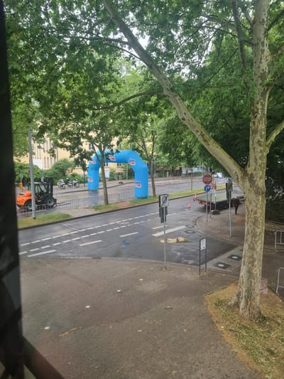 The original image is oriented vertically and rotated 90 degrees clockwise. Described from a corrected perspective looking down onto a street, the scene appears to be set in Saarbrücken, Germany, depicting an area prepared for a public event.
Large, mature trees with distinctive mottled bark and dense green foliage frame the view, suggesting an elevated vantage point. A paved road, appearing wet in areas with puddles and a reflective sheen, runs through the center. It features white dashed lane markings and a possible bicycle symbol on the asphalt. Adjacent to the road is a sidewalk and grassy areas, with yellowish buildings visible in the background on the left.
Two prominent blue inflatable arches, both bearing the "dm" logo, stand on the left side of the road, strongly indicating a sponsored event by the German drugstore chain dm-drogerie markt. Metal crowd control barriers line sections of the street, further supporting the presence of an organized gathering or race. An orange forklift and several motorcycles are parked near the event setup.
Further down the street, a dark car is visible, with a person standing nearby. Various traffic signs are present, including a red octagonal "STOP" sign, a circular blue mandatory right-turn sign, a partially obscured "30" speed limit sign, and a yellow sign with the text "City". Several orange traffic cones are also placed on the pavement.
The scene is illuminated by natural daylight, suggesting it is daytime. The wet ground and soft lighting indicate recent rain or an overcast sky. The overall impression is one of event preparations in an urban environment.