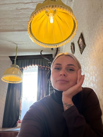 A woman with short blonde hair and a tattoo on her forearm is positioned in the foreground, holding her hand to her face. She is wearing a brown turtleneck shirt. The room appears to be indoors, with a low ceiling covered in a patterned wallpaper. Hanging from the ceiling is a yellow lampshade with fringed edges, illuminated by a visible light bulb. Behind her, a window with a dark frame and curtains allows sunlight to enter the room. Another smaller yellow lampshade is also visible near the window. Two small, framed pictures are affixed to the ceiling. Based on the information provided, the location is Sarapul, Russia.