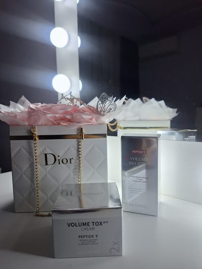 The image presents a still life composition featuring beauty products and decorative items, displayed on a flat surface, possibly a shelf or table. The photograph is oriented vertically, appearing rotated 90 degrees clockwise.

The central subjects are a white "Dior" box with a textured, quilted pattern, and two boxes of "MEDIPEEL+" skincare products. The Dior box is adorned with a delicate gold chain and is accompanied by a pink, ruffled fabric ornament and a silver, butterfly-like metallic decoration.

One MediPeel product box is dark-colored, labeled "PEPTIDE 9 VOLUME BIO TOX pro AMPOM." Visible text on this box includes the ingredients "+ Hydrolyzed Collagen + Ectoin + Hyaluronic Acid" and a partial description: "Advanced nine peptide techo MEDIPEEL offers an ama effect of expert-grade skinomi." The second MediPeel product box is light grey and labeled "VOLUME TOX Pro CREAM PEPTIDE 9 INTENSIVE SKIN CARE WITH 9 PEPTIDES TO ENHANCE ELASTICITY." This box also indicates "2x."

In the background, a reflection of a vanity mirror with several bright, circular lights is visible, casting a glow and suggesting an indoor setting like a dressing area, beauty counter, or salon. The background is predominantly dark. No people or specific events are depicted. The overall arrangement suggests a product display, a gift, or items for personal beauty routine. The location context is Petrich, Bulgaria.