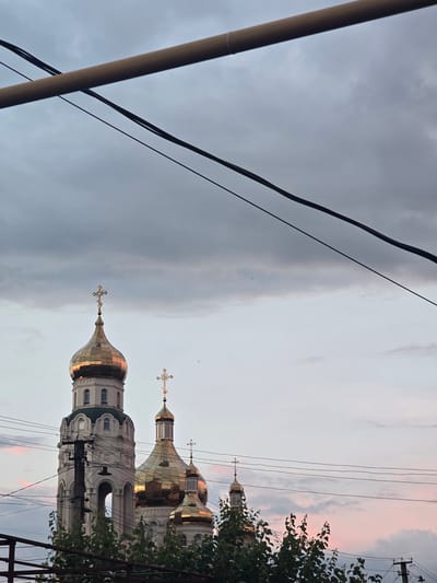 This is a photograph of a church with golden domes and crosses, seen from a low angle. The church has white walls and arched windows. Several utility wires run horizontally across the frame, obscuring parts of the sky and the upper sections of the church. A tree with green leaves is visible on the right side of the image. The sky is overcast with dramatic clouds, tinged with pink and orange hues, suggesting either sunrise or sunset. The overall mood is somewhat dramatic due to the lighting and cloudy sky. The image is oriented vertically, with the sky appearing at the top. There is no visible text in the image.