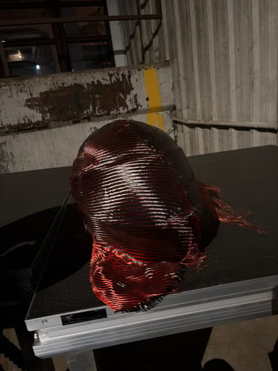 A close-up shot captures a dark, textured object with a red and black woven pattern, possibly a fabric or a piece of art, resting on a black, flat surface. The object appears to be spherical or dome-shaped at the top and tapers down, with loose red threads visible at the bottom. The background consists of a ribbed, grey metal wall with vertical lines and a weathered concrete surface with patches of rust-colored discoloration and a yellow stripe. A dark metal railing is also visible in the upper left portion of the image. The scene is dimly lit, with highlights reflecting off the woven material. The location is an outdoor urban environment, possibly an industrial area or an alleyway in Rotterdam, Netherlands, suggested by the building materials and urban setting.