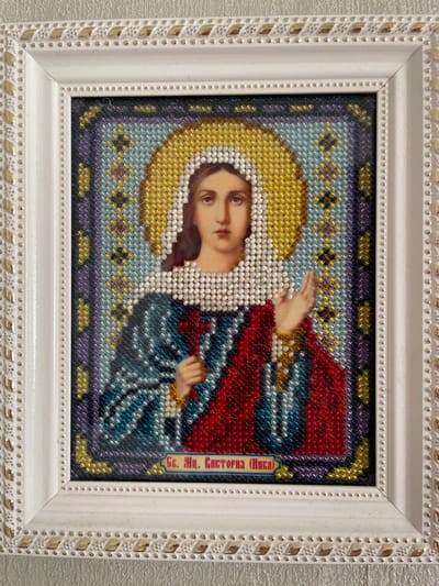 This is a framed image of a religious icon, likely a depiction of the Virgin Mary, created using bead embroidery. The central figure is a woman with a halo, rendered in shades of white and yellow beads. Her face is detailed with brown and pink beads, and she is looking slightly to the left with a calm expression. She wears a red garment over a blue and teal robe. Her right hand is raised, and her left hand is visible near her waist.
The background of the icon is composed of intricate patterns of beads in shades of dark blue, purple, light blue, and gold. There are star-like motifs in gold and purple within the background. On the right side of the icon, there is a vertical band of text in Cyrillic script, embroidered in red and yellow beads. The text appears to read "Св. Мц. Виктория (Икона)" which translates to "St. Martyr Victoria (Icon)".
The beadwork is framed by a white, textured frame with a decorative border. The border of the frame features a repeating pattern of small gold beads and larger gold-colored rectangular elements. The overall presentation suggests a devotional or artistic piece. The context of Brest, Belarus, implies this is likely an item of religious or cultural significance within that region.