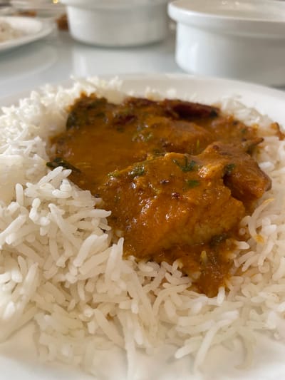 A plate of food is displayed, featuring white rice and a portion of chicken curry. The dish is served on a white plate, and there are indications of additional tableware visible in the blurred background, suggesting a dining setting. The location is presumed to be Bengaluru, India, based on the presence of Indian cuisine.