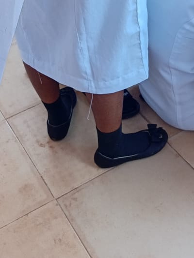 The scene in Ejule, Nigeria, displays the lower extremities of at least two individuals on a tiled floor. One person, positioned on the left, shows bare lower legs with a dark skin tone, wearing black ankle socks and plain dark flat shoes. A light-colored garment, possibly a skirt or trousers, covers the upper portion of their legs, with a loose white thread hanging from its hem. To the right, part of a second individual is visible, also wearing a light-colored garment, black ankle socks, and dark flat shoes, with the right shoe featuring a small decorative bow. The floor is composed of light-beige square tiles with dark grout lines, exhibiting minor smudges and discoloration.