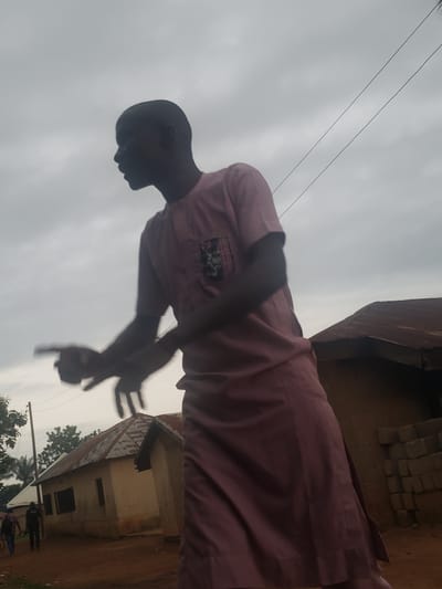 In Gumel, Nigeria, under a cloudy sky, a person stands outdoors, captured in silhouette against the overcast atmosphere. They are wearing a pink, ankle-length traditional garment with short sleeves and a detailed pattern on the chest pocket. Their head is turned to the left, with facial features obscured by shadow. Their right arm is extended forward, with their hand pointing, while their left arm is bent at the elbow and also extended. Behind the person, two simple, single-story structures with corrugated metal roofs are visible. The ground is reddish-brown earth. In the distance, another building and trees can be seen, along with a utility pole. The image appears to be taken from a low angle, emphasizing the figure against the sky.