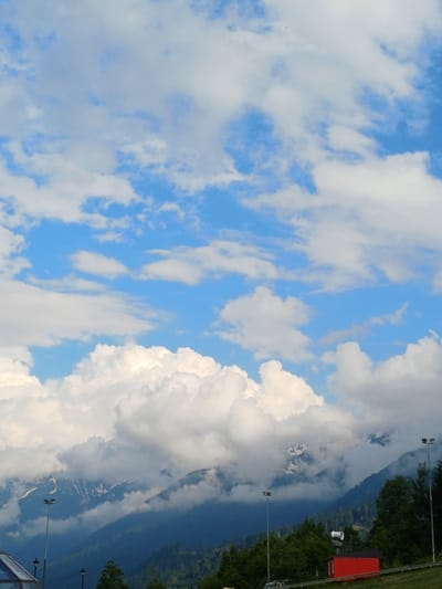 This image shows a mountainous landscape in Estosadok, Russia. The sky is filled with fluffy white clouds against a bright blue backdrop. In the mid-ground, majestic mountains rise, their peaks partially obscured by clouds and snow. The lower slopes are covered with dense green trees. In the foreground, there is a red building with a white conical structure on top, possibly a snow cannon or a weather station. Several lampposts with spotlights are visible, suggesting a ski resort or a similar recreational area. The overall scene depicts a natural, scenic environment with man-made structures integrated into the landscape.