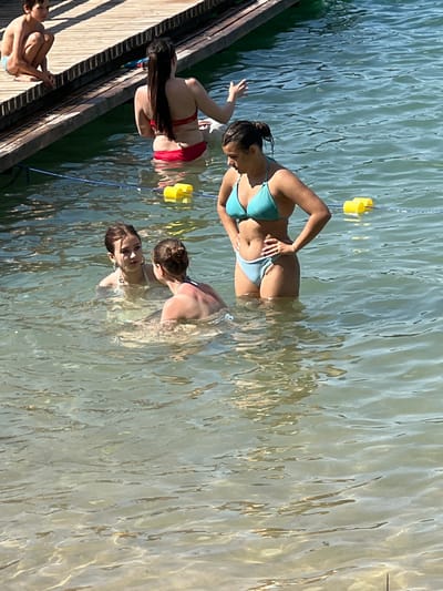 In Vilnius, Lithuania, a group of young people are enjoying a sunny day at a lake. The image captures a candid moment as they stand in the clear, shimmering water near a wooden dock. The water is shallow enough for them to stand comfortably, and the sunlight creates a dappled effect on the surface.
Several individuals are visible in the water, all wearing swimwear. In the foreground, a person in a red bikini is partially submerged, with their long dark hair cascading around them. To their left and slightly behind, another person in a light blue bikini is looking towards the viewer with a smile. Further into the water, two more individuals are visible, one in the background with their back to the camera and another in the mid-ground facing away from the camera. A young boy is seated on the wooden dock in the lower left corner of the frame, appearing relaxed.
The scene suggests a leisurely summer day, with the bright sunlight and clear water indicating fair weather. The wooden dock suggests a recreational area, possibly a beach or a designated swimming spot. The overall mood is one of carefree enjoyment and relaxation. There is no visible text in the image.