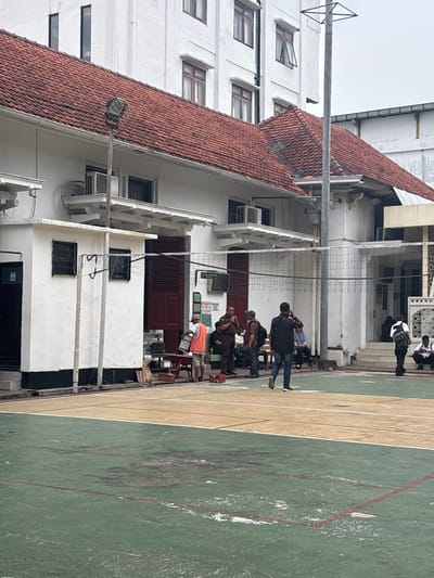 A group gathers on a green and beige painted court next to a white building with a red tile roof. Some individuals are seated, and others stand. A volleyball net stretches across the center. The building has multiple windows and air conditioning units. In the background, more people stand near an archway, and the sky is overcast.